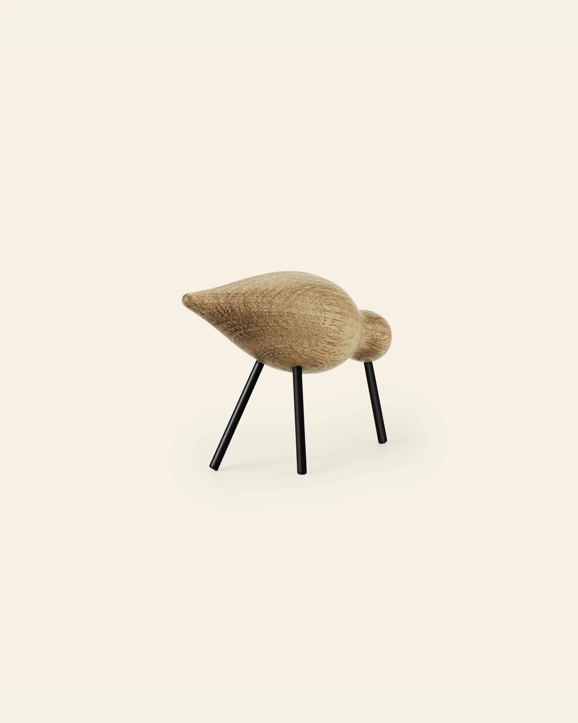 The Normann Copenhagen Shorebird Medium Oak is a minimalist oak wood bird sculpture by Normann Copenhagen, featuring a smooth rounded body and head on four thin black metal legs, shown against a plain light background.
