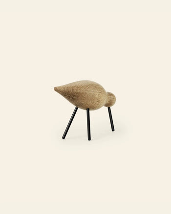The Normann Copenhagen Shorebird Medium Oak is a minimalist oak wood bird sculpture by Normann Copenhagen, featuring a smooth rounded body and head on four thin black metal legs, shown against a plain light background.