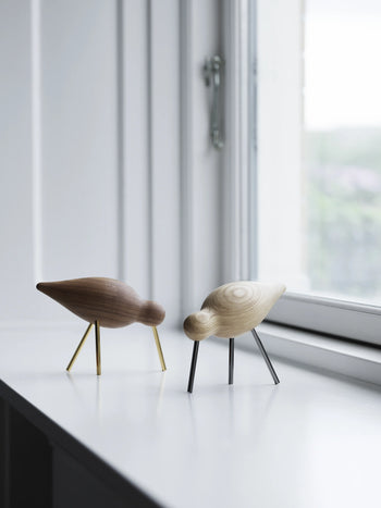 A Normann Copenhagen Shorebird Medium Oak sculpture stands on a white windowsill, its oak body and black legs bathed in soft natural light filtering through the window.