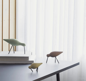 The Normann Copenhagen Shorebird Medium Oak by Normann Copenhagen, with slender legs and minimalist form, is displayed on a gray table. Two stand directly on the surface while one rests on stacked white books amid sheer curtains and wood panels.