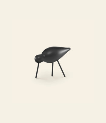 The Normann Copenhagen Shorebird Medium Oak is a minimalist black sculpture with a rounded body and head, pointed beak, and thin oak legs, standing elegantly against a plain light background.