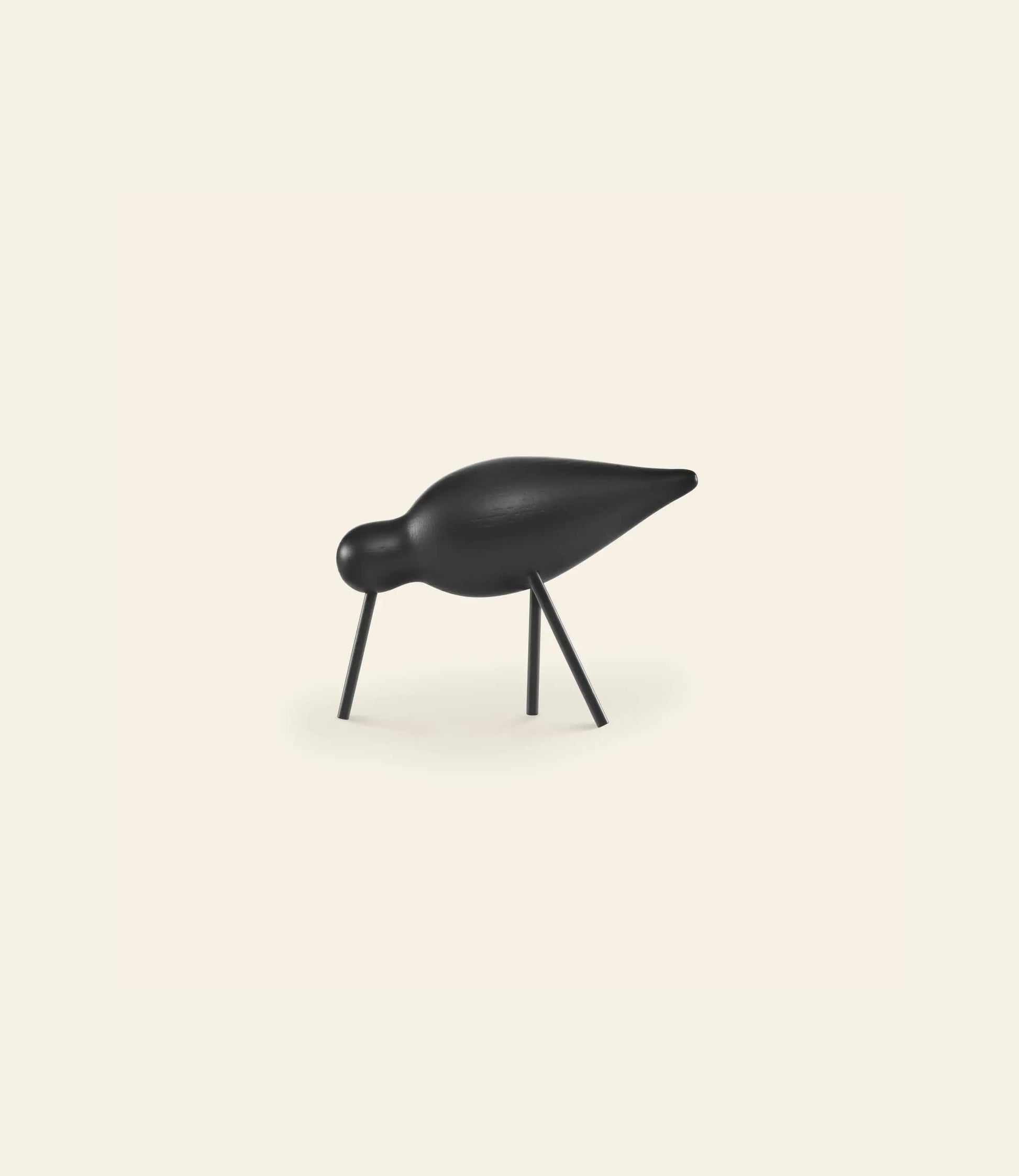 A minimalist black Normann Copenhagen Shorebird Medium Oak sculpture features a rounded oak body, small head, pointed beak, and thin legs poised as if pecking the ground against a plain light background.
