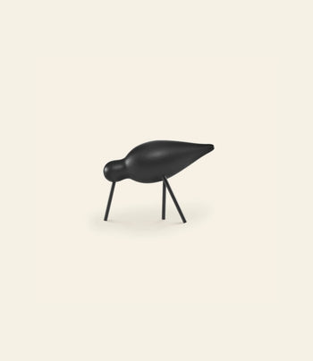 A minimalist black Normann Copenhagen Shorebird Medium Oak sculpture features a rounded oak body, small head, pointed beak, and thin legs poised as if pecking the ground against a plain light background.