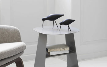 A modern gray side table holds a Normann Copenhagen Shorebird Medium Oak sculpture and a closed book on its lower shelf, next to a light gray upholstered chair in a bright, white minimalist room.