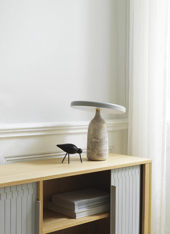 A minimal scene with an oak wood cabinet, a round ceramic lamp, and the Normann Copenhagen Shorebird Medium Oak figurine by Normann Copenhagen, alongside stacked magazines against a white wall with sheer curtains.