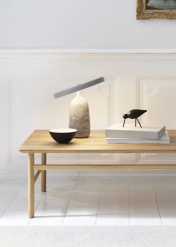 A minimalist oak wood bench displays a stone lamp, a black bowl, and decorative books topped with the Normann Copenhagen Shorebird Medium Oak sculpture by Normann Copenhagen, set against a white paneled wall.