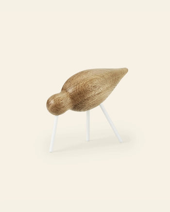 The Normann Copenhagen Shorebird Medium Oak is a minimalist oak figurine with a smooth, rounded body and head, standing on three slender white legs against a light background.