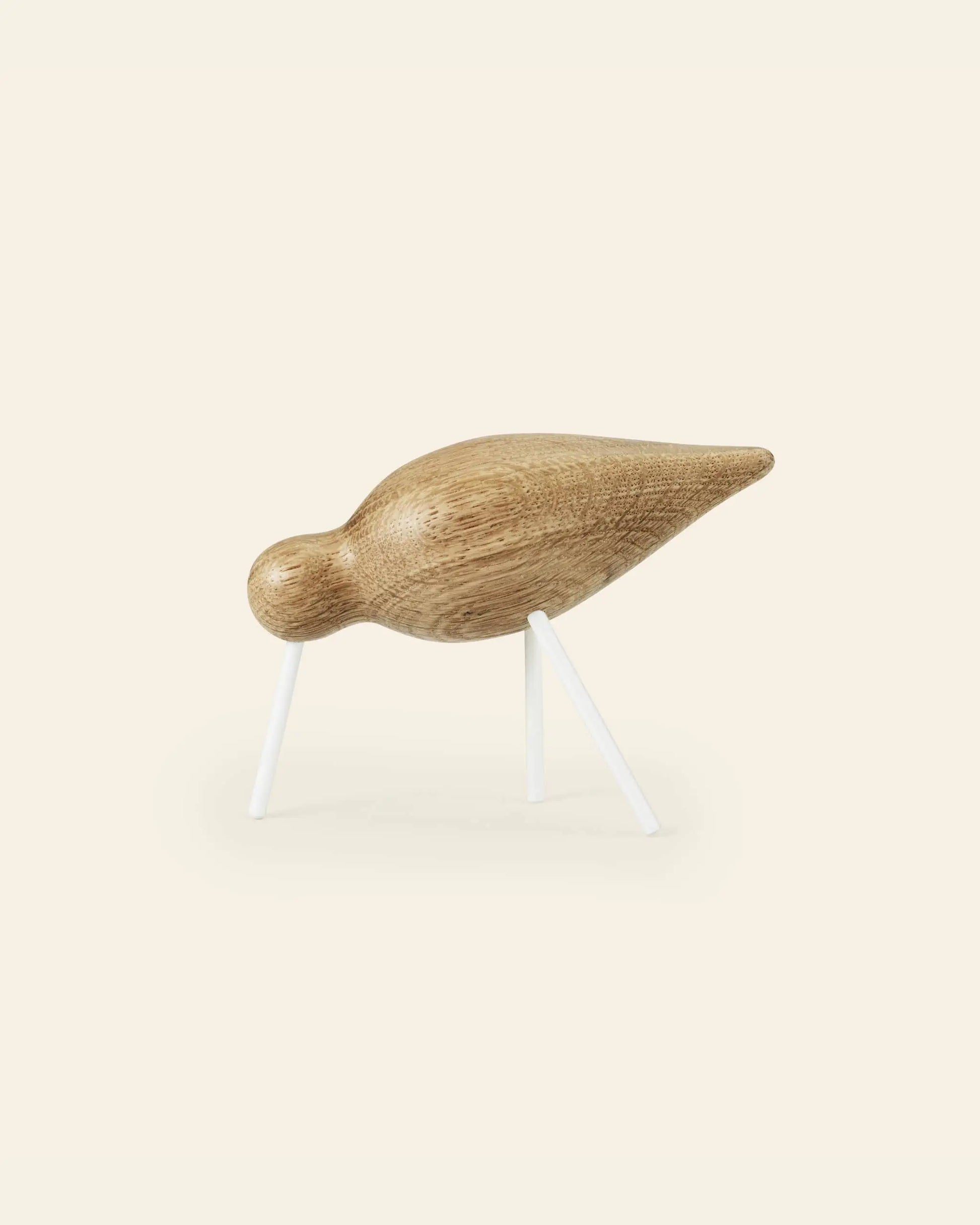 The Normann Copenhagen Shorebird Medium Oak features a minimalist, rounded oak body and head on slim white legs, inspired by bird forms from Normann Copenhagen, set against a light beige background.