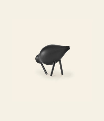 A minimalist black sculpture, reminiscent of the Normann Copenhagen Shorebird Small Oak by Normann Copenhagen, features a stylized animal form with a rounded body and four angled legs, set against an off-white background.