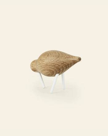 The Normann Copenhagen Shorebird Small Oak is a minimalist wooden bird sculpture with a rounded oak body, pointed beak, and two white legs, from the brand Normann Copenhagen, displayed on a light background.