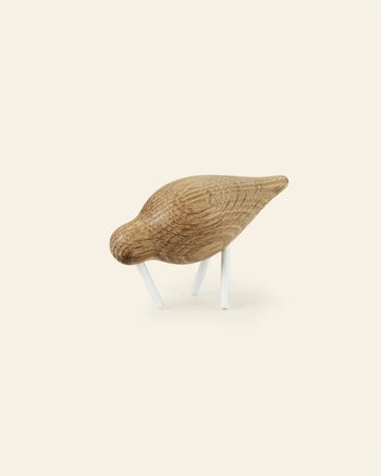 The Normann Copenhagen Shorebird Small Oak features a minimalist oak wood design with a rounded body, pointed tail, and white legs, appearing to peck downward against a plain light background.