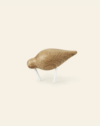 The Normann Copenhagen Shorebird Small Oak, a minimalist oak wood bird sculpture with a smooth, rounded body and two white legs, stands as if pecking against a plain light background.