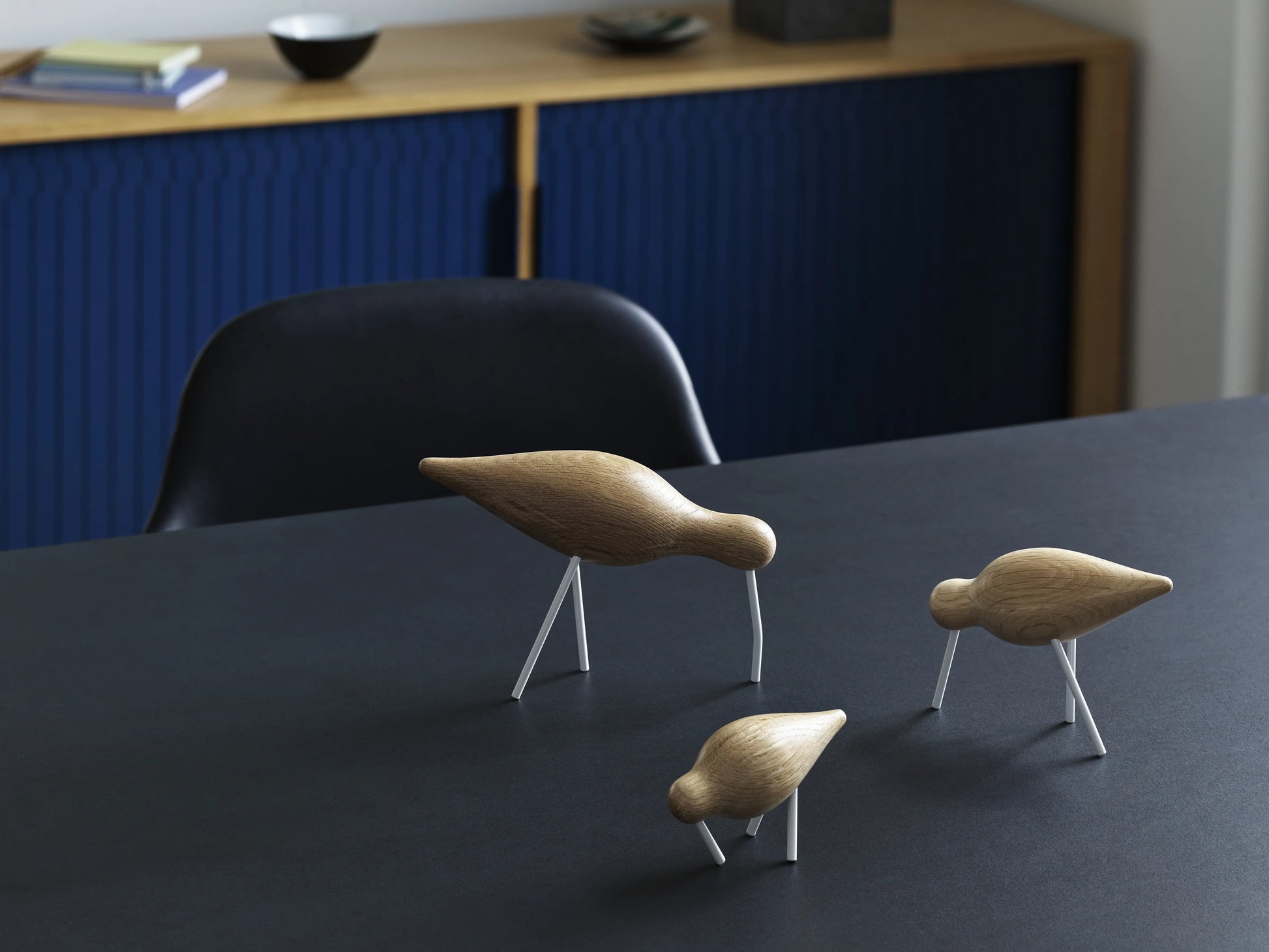 Three minimalist Normann Copenhagen Shorebird Small Oak figurines stand on a dark table. In the background, a black chair and a blue cabinet with oak trim display small decorative items.