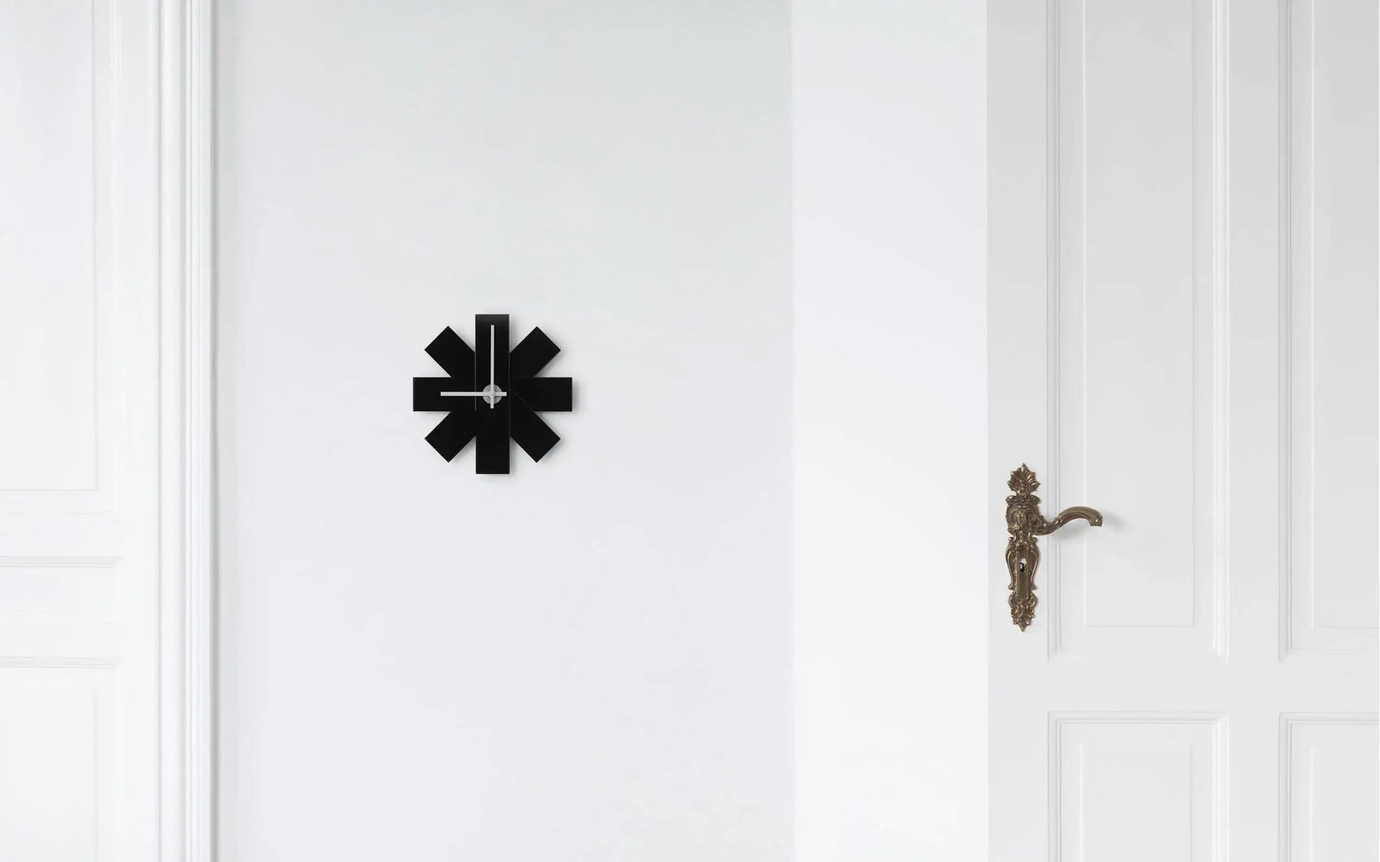 The Normann Copenhagen Watch Me Wall Clock by Normann Copenhagen features minimalist, abstract, angular shapes in black—an ideal modern decor piece for a plain white wall next to a white door with an ornate brass handle.