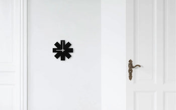 The Normann Copenhagen Watch Me Wall Clock by Normann Copenhagen features minimalist, abstract, angular shapes in black—an ideal modern decor piece for a plain white wall next to a white door with an ornate brass handle.
