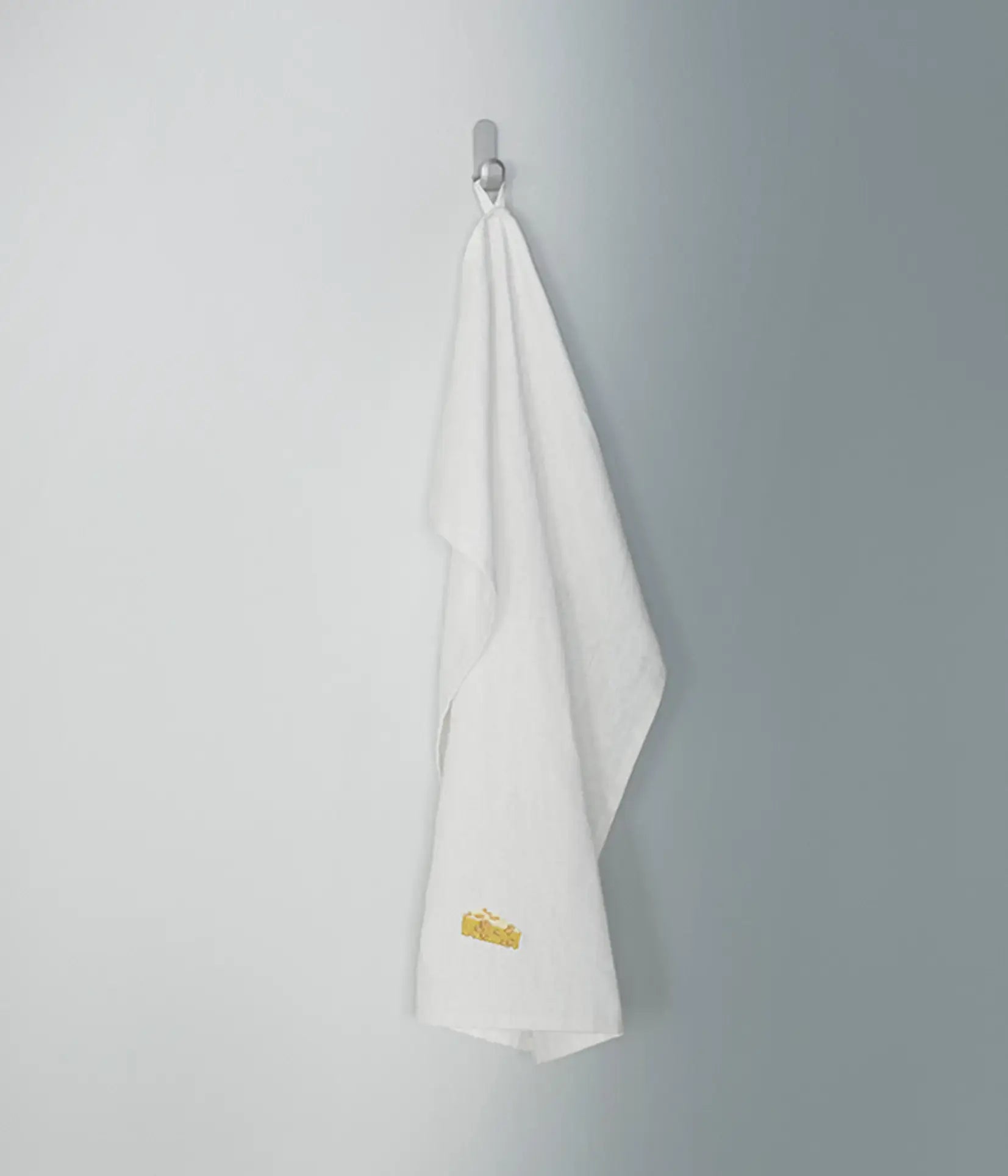 The Normann Copenhagen Yummy Tea Towel, crafted from white linen and cotton with a subtle yellow embroidered accent, hangs from a silver hook on a smooth, light gray wall.