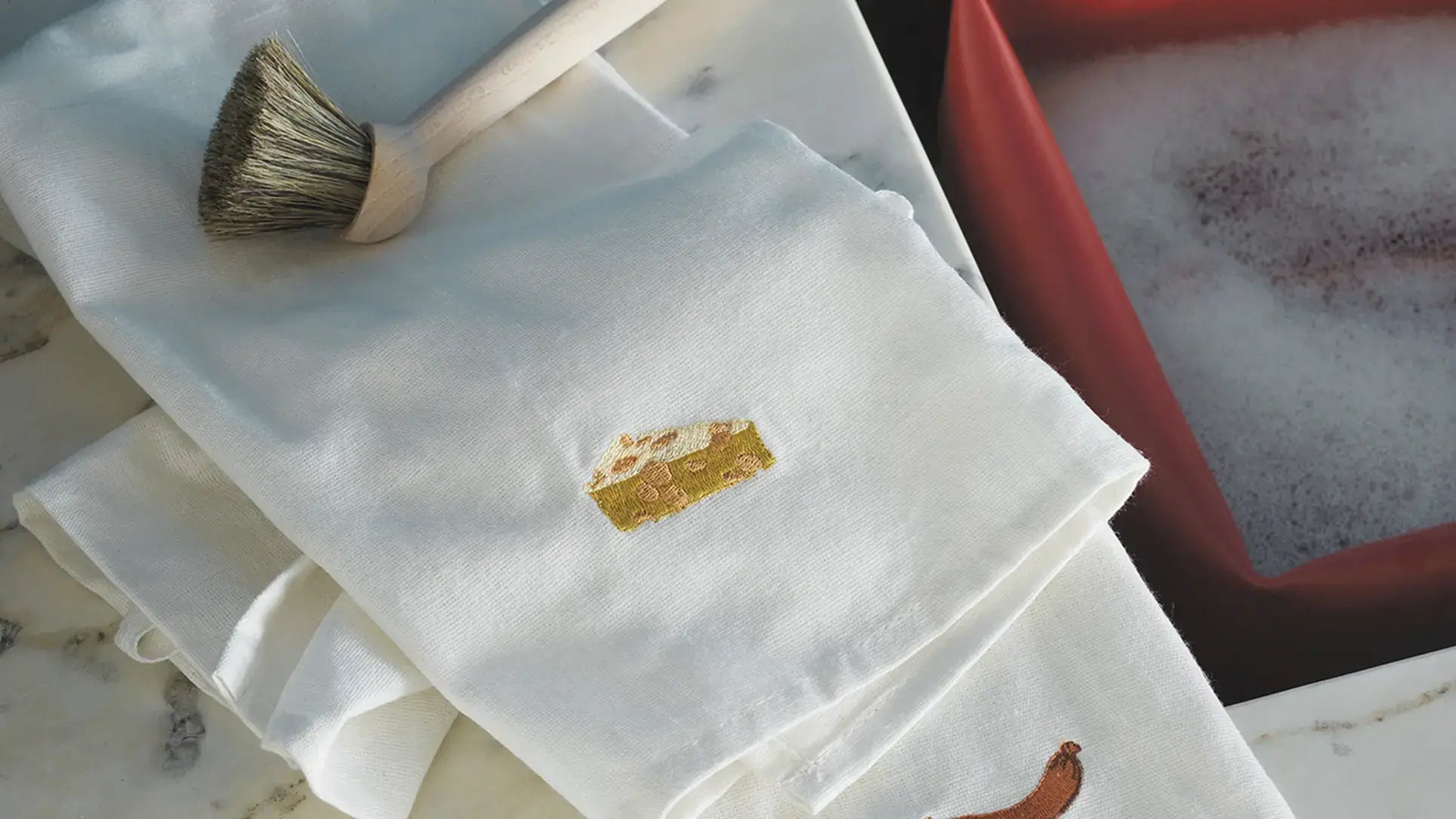 The Normann Copenhagen Yummy Tea Towel in white, OEKO-TEX certified and featuring an embroidered cheese wedge, is folded on marble beside a natural bristle brush and a red container of soapy water.