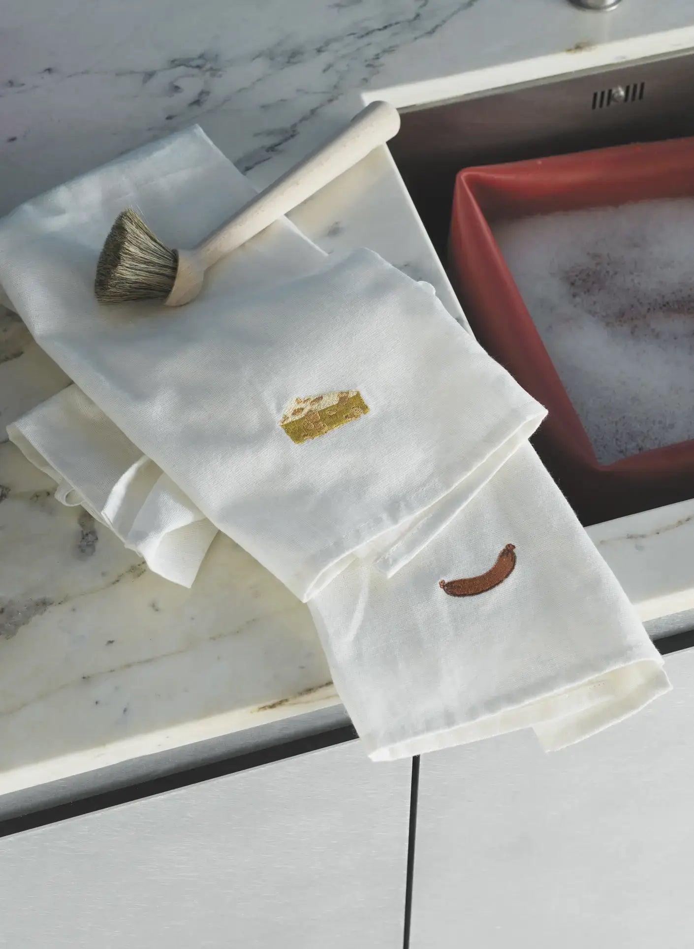 Two Normann Copenhagen Yummy Tea Towels by Normann Copenhagen, featuring embroidered cheese and sausage designs, rest on a marble countertop near a dish brush and a red sink with soapy water.