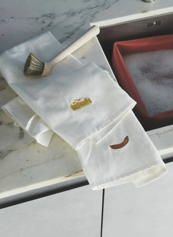 Two Normann Copenhagen Yummy Tea Towels by Normann Copenhagen, featuring embroidered cheese and sausage designs, rest on a marble countertop near a dish brush and a red sink with soapy water.