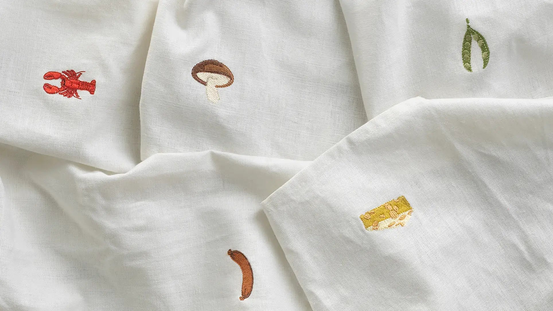 The Normann Copenhagen Yummy Tea Towel features white linen-cotton fabric with embroidered motifs—red lobster, brown mushroom, green peas, cheese block, and sausage. OEKO-TEX certified for a whimsical and safe kitchen accent.