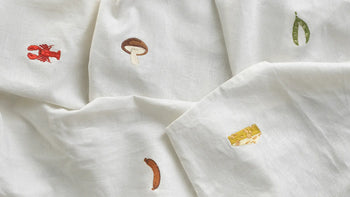 The Normann Copenhagen Yummy Tea Towel features white linen-cotton fabric with embroidered motifs—red lobster, brown mushroom, green peas, cheese block, and sausage. OEKO-TEX certified for a whimsical and safe kitchen accent.