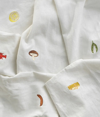 The Normann Copenhagen Yummy Tea Towel features white linen-cotton fabric with playful embroidered food motifs—lemon, pea pod, mushroom, sausage, cheese, and chili—perfect for adding charm to your kitchen.