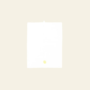 A plain white sheet of paper is set against a light beige background, featuring a small yellow circular sticker near the bottom center—echoing the clean, fresh design of the Normann Copenhagen Yummy Tea Towel by Normann Copenhagen.