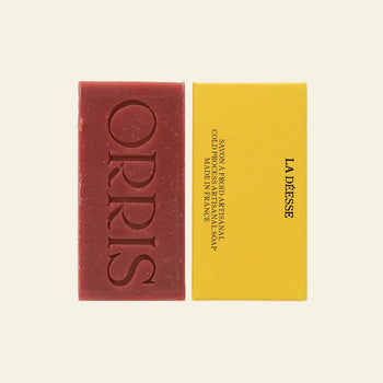 A rectangular red ORRIS La Déesse Botanical Soap bar, with ORRIS embossed on it and containing Ayurvedic Manjistha, sits beside a yellow box labeled La Déesse in black text. The scene is set against an off-white background.