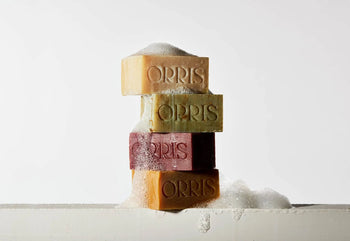 Four stacked ORRIS Le Nomade artisanal soap bars in assorted colors are topped and surrounded by white bubbles against a plain white background. The scene evokes subtle cedarwood and turmeric notes.