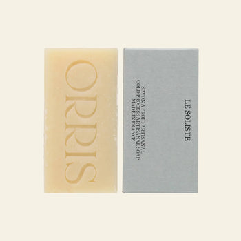 A rectangular beige ORRIS Le Soliste artisanal soap bar, embossed with ORRIS, rests beside its light grey packaging labeled LE SOLISTE. The cruelty-free bar is set against a plain light background.