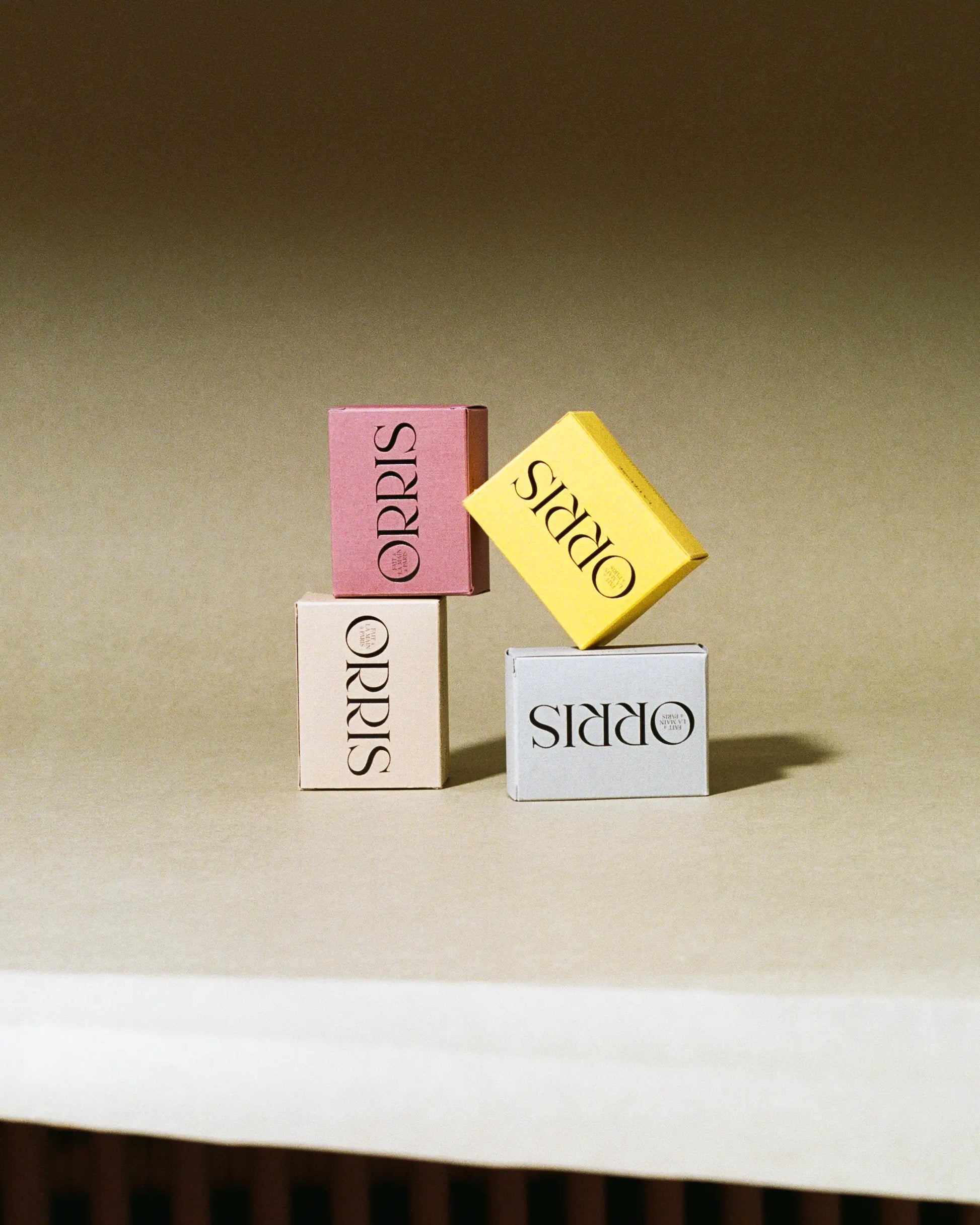 Three small artisanal soap boxes from ORRIS, including Le Soliste in yellow, are stacked on a beige surface. The yellow box highlights postbiotic fermented rice water for a cruelty-free cleanse.