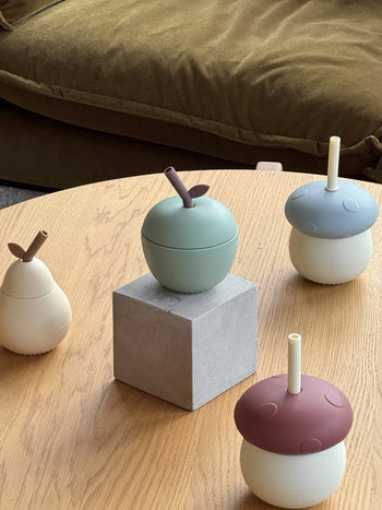 Four pastel silicone fruit- and nut-shaped cups with straws sit on a wooden table; the green OYOY Living Design Apple Cup is on a gray block, with pear and acorn designs arranged nearby.