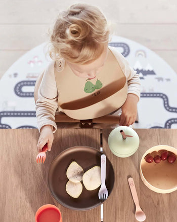 A young child with curly blonde hair, wearing a beige bib with a pear design, sits at a table with sliced pears, grapes, utensils, and the OYOY Living Design Apple Cup - Green by OYOY Living Design, viewed from above.