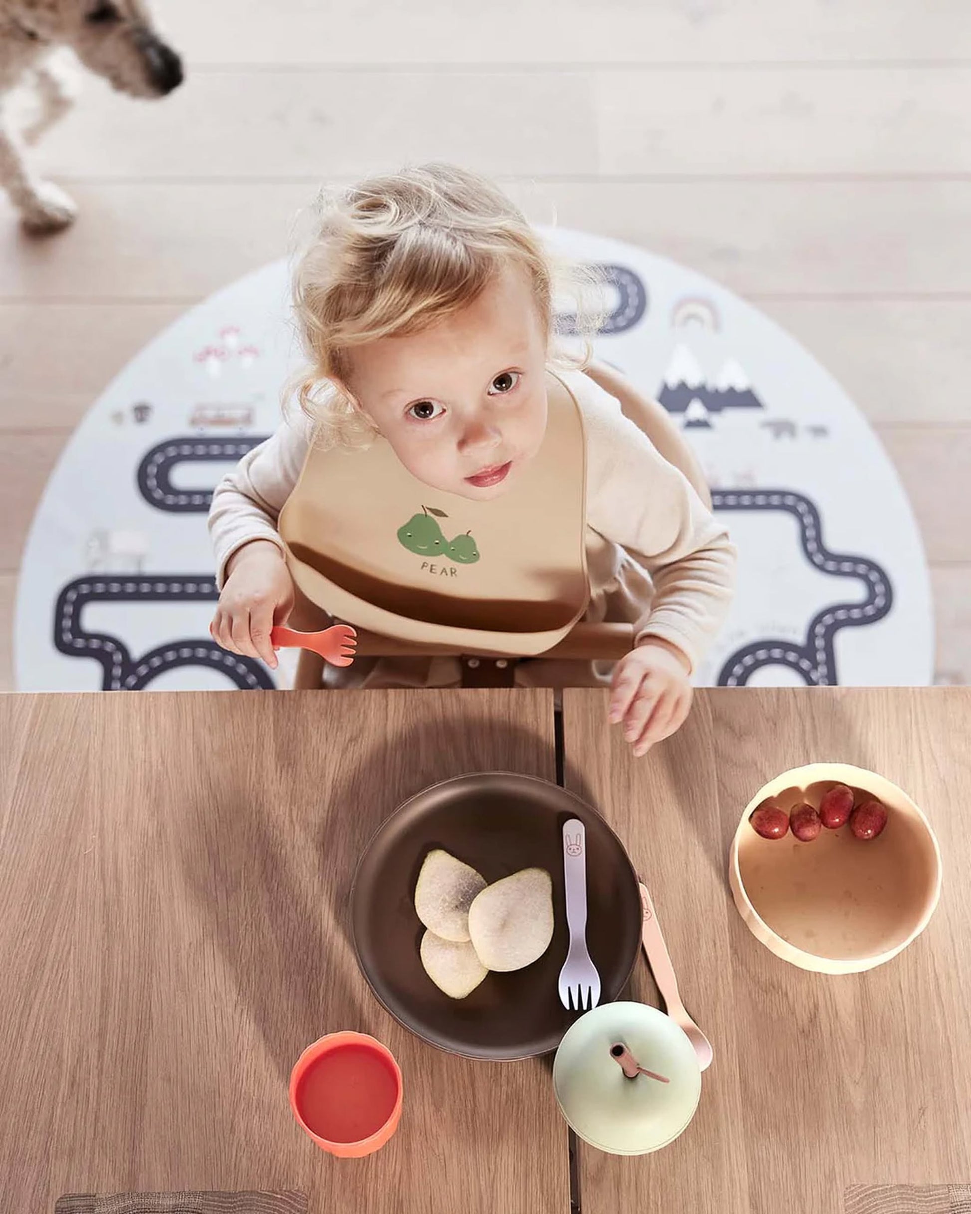 A young blond-haired child sits at a wooden table with bowls of pears and grapes, an OYOY Living Design Apple Cup - Green, a fork, and an apple-shaped container. A dogs paw appears in the background.