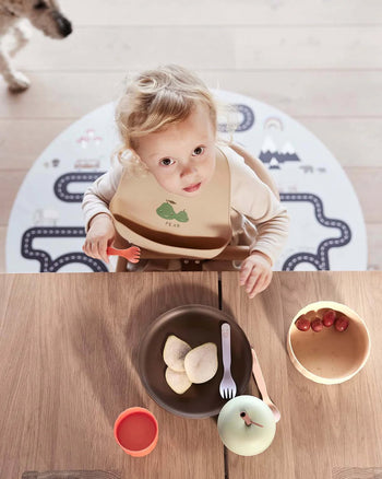 A young blond-haired child sits at a wooden table with bowls of pears and grapes, an OYOY Living Design Apple Cup - Green, a fork, and an apple-shaped container. A dogs paw appears in the background.