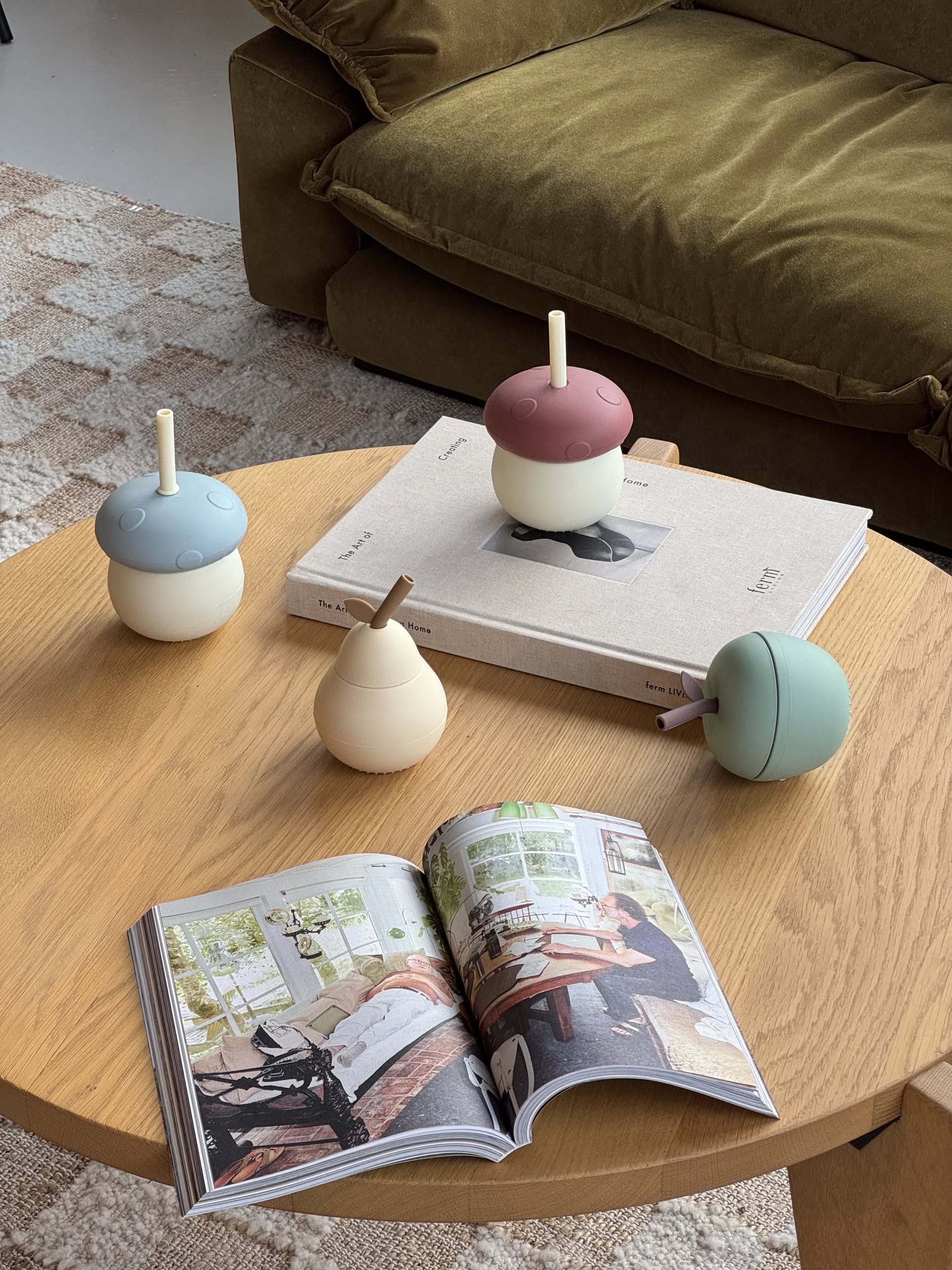 Four acorn-shaped silicone drink cups, including the green OYOY Living Design Apple Cup, rest on a round wooden coffee table with two books. A brown sofa and textured rug serve as the backdrop.