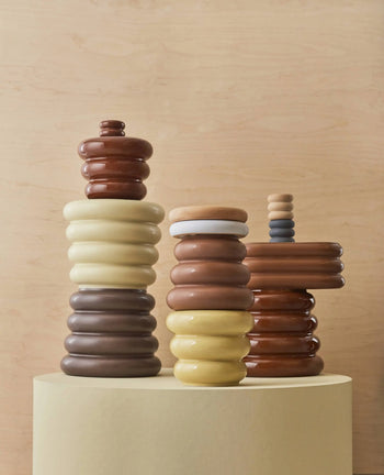 Four glossy, rounded OYOY Living Design Awa Pots (Large) in neutral brown, beige, cream, yellow, and gray sit atop a light circular surface with a light wooden background.