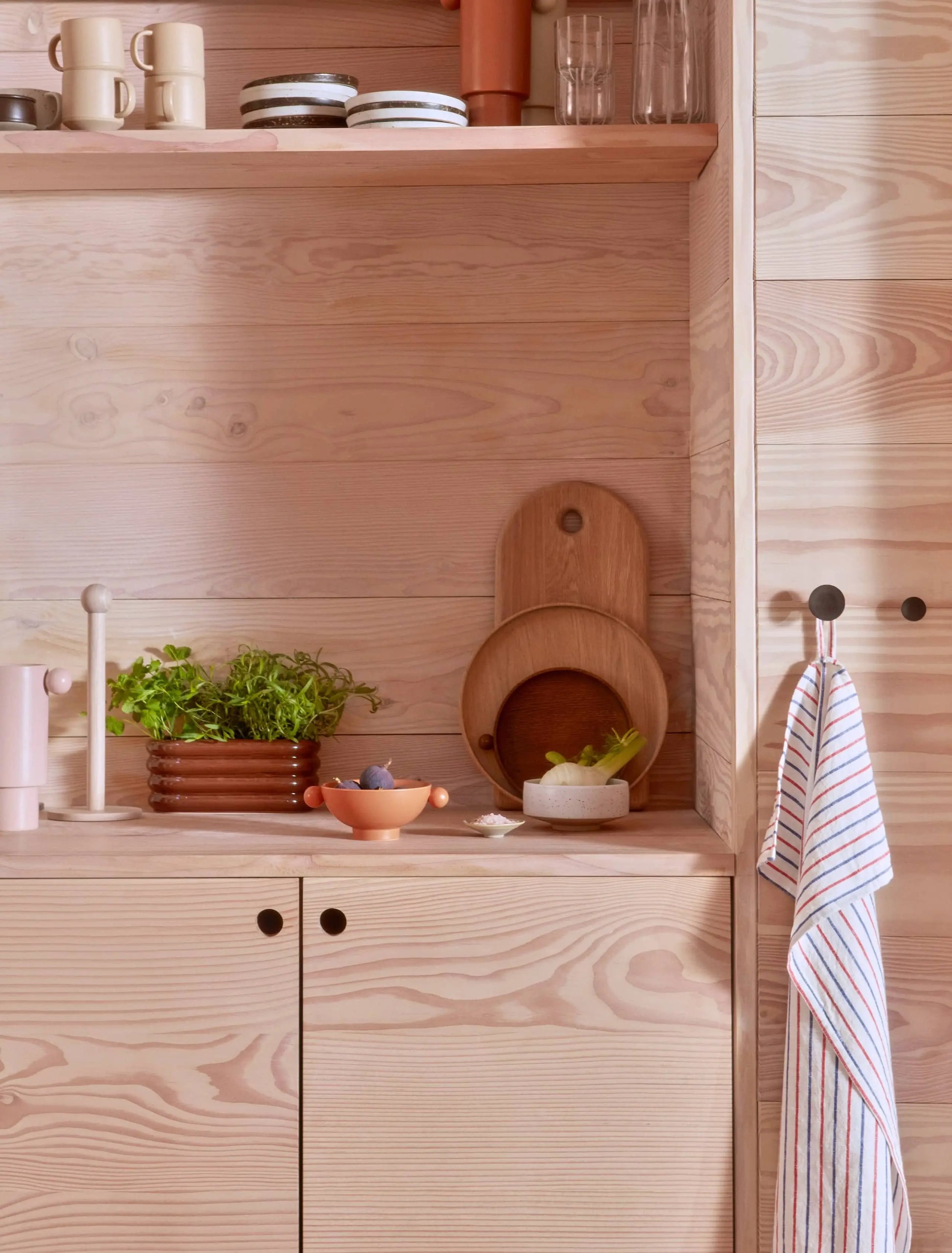 Light wood kitchen with shelves, plates, bowls, plants, cutting boards, and the OYOY Living Design Awa Pot, Long. Minimalist and organized with natural tones, earthenware touches by OYOY Living Design, and a striped towel on a knob.