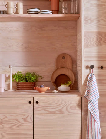 Light wood kitchen with shelves, plates, bowls, plants, cutting boards, and the OYOY Living Design Awa Pot, Long. Minimalist and organized with natural tones, earthenware touches by OYOY Living Design, and a striped towel on a knob.
