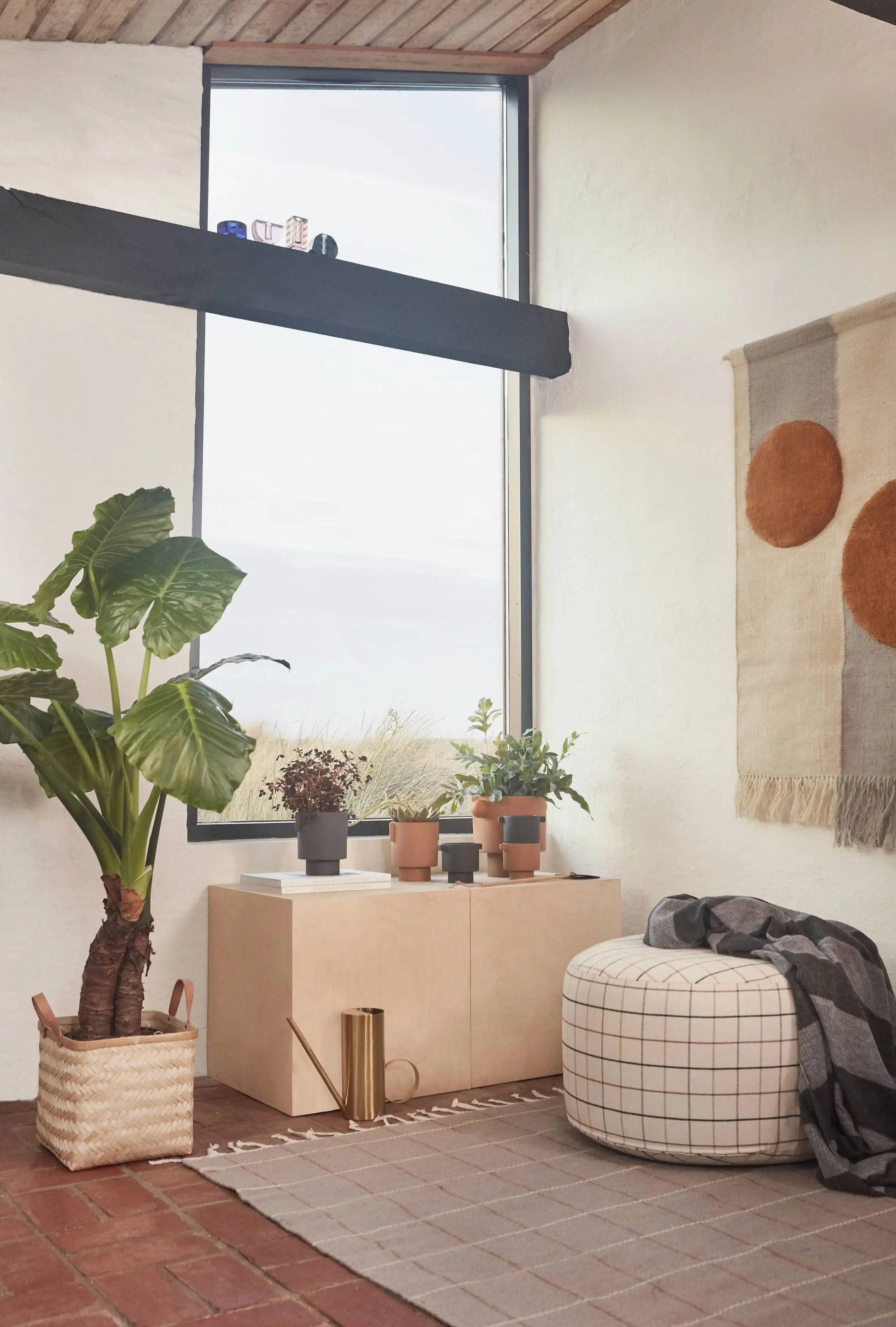 A cozy, modern room with a large plant, potted plants on a cabinet, gold watering can, an OYOY Living Design Grid Rug, round plaid ottoman with gray blanket, and wall hanging with two brown circles.