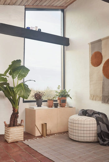 A cozy, modern room with a large plant, potted plants on a cabinet, gold watering can, an OYOY Living Design Grid Rug, round plaid ottoman with gray blanket, and wall hanging with two brown circles.