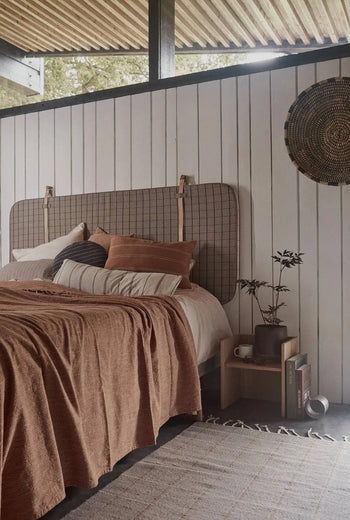 A cozy bedroom features rust bedding, striped pillows, and a textured headboard. The OYOY Living Design Grid Rug anchors the space, while a nightstand with books and a plant sits under woven wall decor.