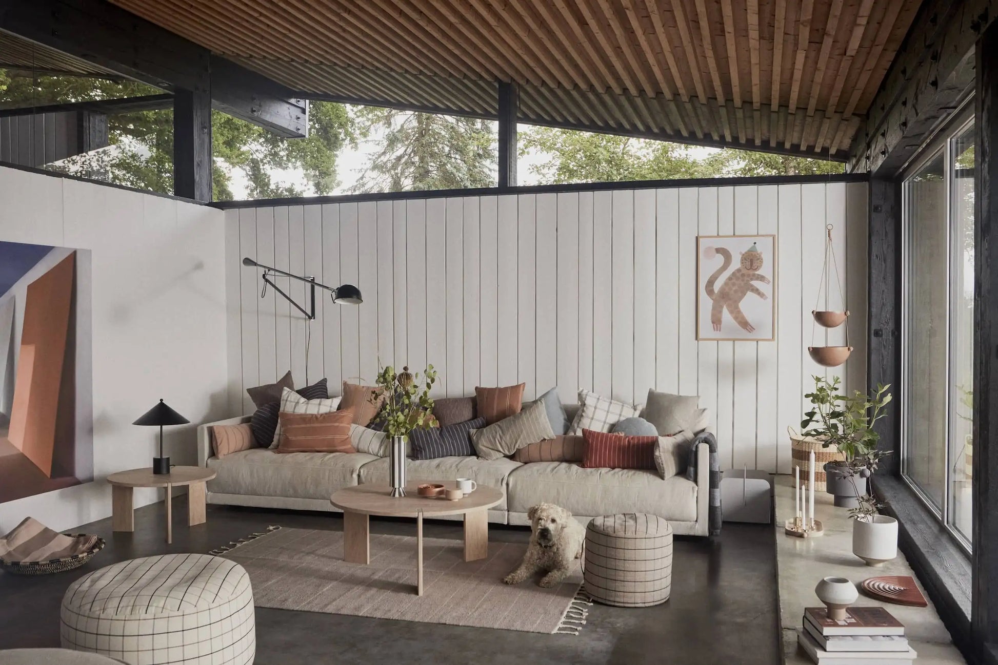 A modern living room with a light sofa, cushions, round coffee table, poufs, indoor plants, and a small dog on the OYOY Living Design Grid Rug. Large windows, wood-paneled ceiling, and white shiplap walls complete the space.