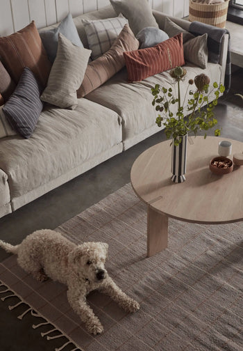 A curly-haired dog rests on the OYOY Living Design Grid Rug, featuring graphic squares, placed by a beige sofa with assorted pillows; a round wooden coffee table holding a floral vase and small bowls is nearby.