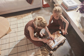 Two young children with blonde hair in buns, dressed in brown, sit on an OYOY Living Design Grid Rug and play with a wooden geometric puzzle set in a bright, cozy living room.