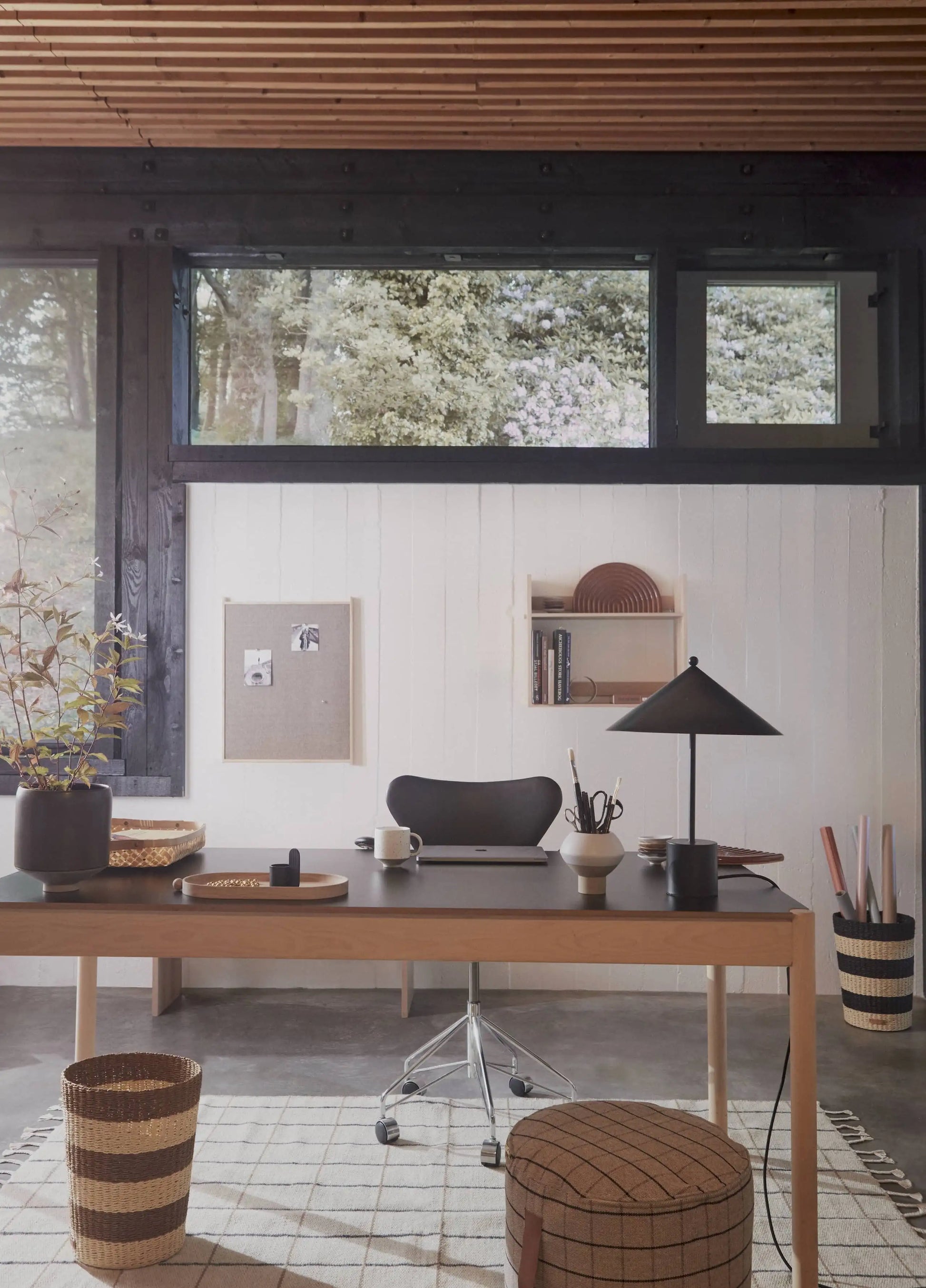 A minimalist home office with a wooden desk, black chair, table lamp, plant, and organized supplies is bathed in sunlight from large windows above, featuring the OYOY Living Design Grid Rug with bold graphic squares beneath the workspace.