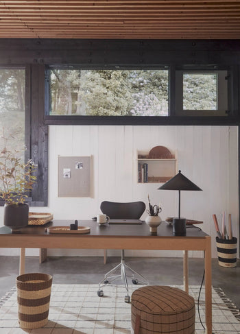 A minimalist home office with a wooden desk, black chair, table lamp, plant, and organized supplies is bathed in sunlight from large windows above, featuring the OYOY Living Design Grid Rug with bold graphic squares beneath the workspace.