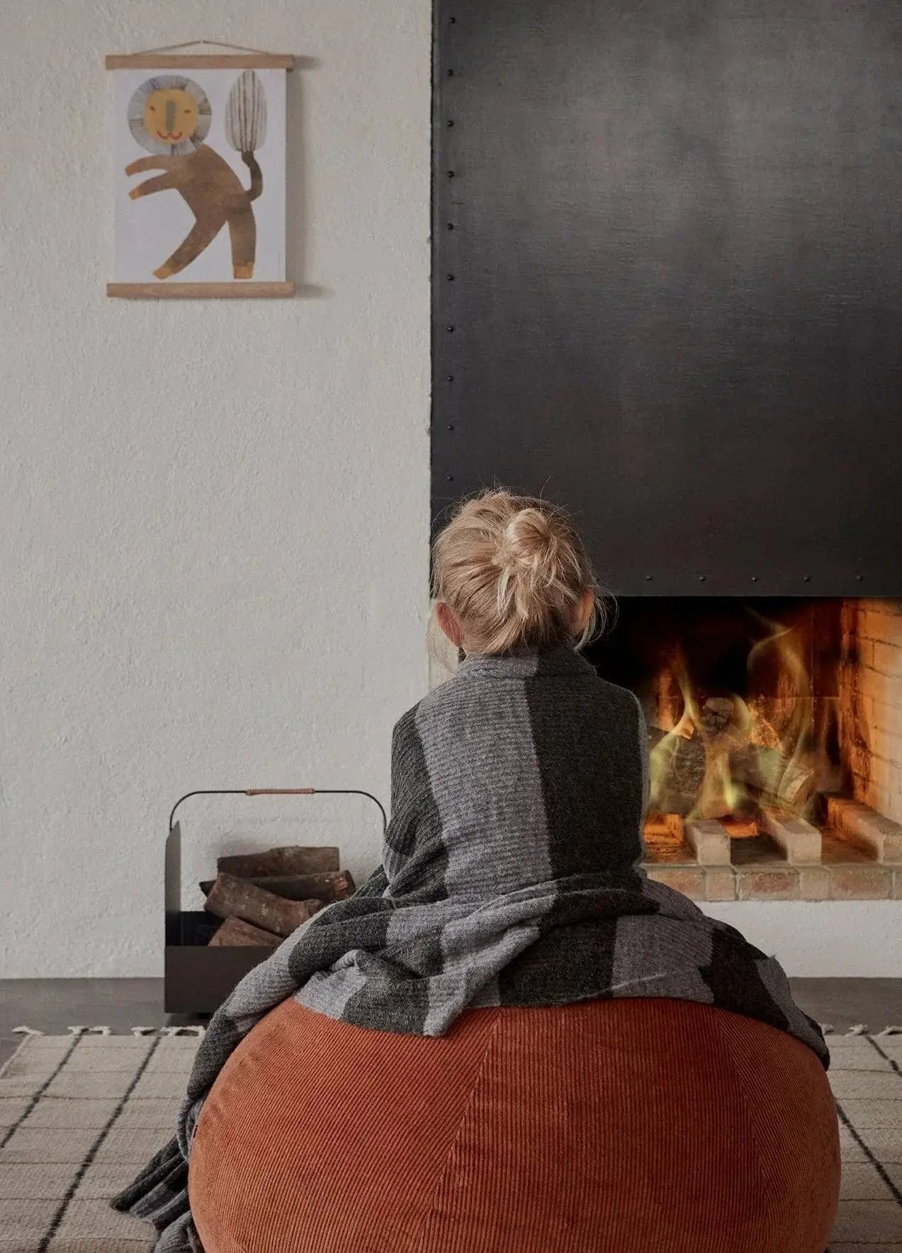A blonde-haired child sits on a brown pouf by a lit fireplace, wrapped in a gray reversible blanket. Firewood is stacked nearby, and playful lion art hangs above the OYOY Living Design Grid Rug on the white wall.