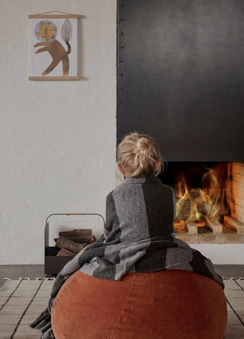 A blonde-haired child sits on a brown pouf by a lit fireplace, wrapped in a gray reversible blanket. Firewood is stacked nearby, and playful lion art hangs above the OYOY Living Design Grid Rug on the white wall.