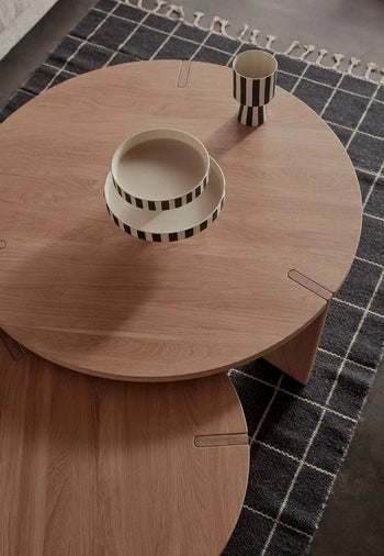 A round wooden coffee table with two black-and-white striped ceramic bowls and a matching cup sits on the OYOY Living Design Grid Rug from OYOY Living Design, featuring graphic squares and tasseled edges.
